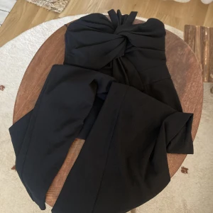 Svart jumpsuit med knut framtil  - Elegant jumpsuit från ginatricot i storlek 38. Smickrande passform. Säljes pga att den inte passar längre. Fin i skick. Nypris 699kr.