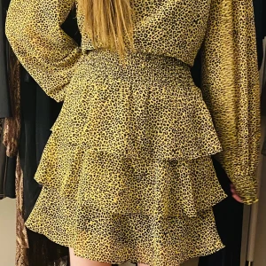 Leopard mönster klänning med volang - Söt och läcker klänning med volanger. Senap klänning med pyttesmå svarta prickar som formar ett leopard mönster. Långärmad. Hög urringning.