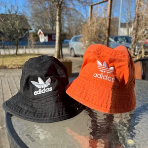Adidas bucket hat  - Säljer två fina adidas bucket hats. Den ena svart och den andra orange. De har olika form vilket syns på de två sista bilderna. Köpta från Zalando och är i storlek M/L. De är sparsamt använda och i väldigt bra skick. Säljs för 200kr för båda, eller 100kr/st.