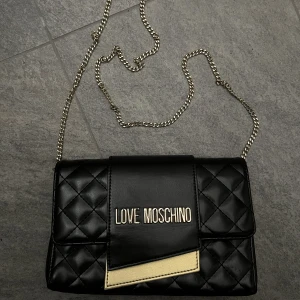 love moschino väska - En love moschino väska i svart med guld detaljer och en guldkedja. I väldigt fint skick få gånger använd. Inköpt för 2 år sedan och just nu ligger den bara i garderoben. Tygpåsen till väskan kommer med också. Nypris 1400kr 