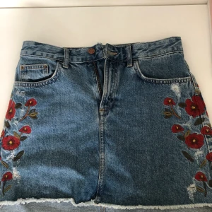 Jeanskjol med broderi detaljer - En jeanskjol lite kortare fram och lite längre där bak. Storlek S men känns lite stor i storleken. Den har röda broderade blommor på sidorna