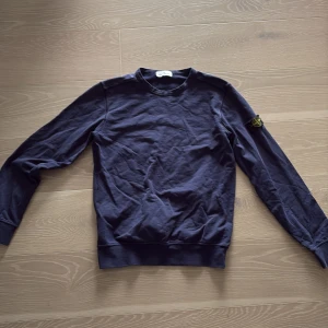 Mörkblå tröja från Stone Island Junior - Säljer en snygg mörkblå tröja från Stone Island Junior. Tröjan har långa ärmar och en rund halsringning. Den ikoniska logotypen sitter på ärmen. Perfekt för en stilren look.