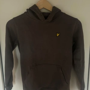 Brun hoodie från Lyle & Scott - Säljer en snygg brun hoodie från Lyle & Scott med deras ikoniska logga på bröstet. Perfekt för en stilren och bekväm look. Hoodien har en klassisk känguruficka framtill och långa ärmar.