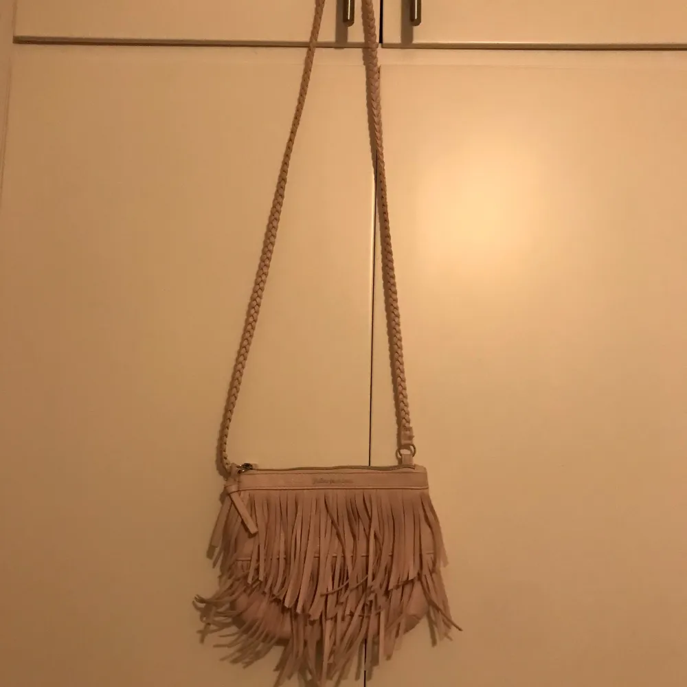 Snygg beige axelväska med coola fransar och flätad axelrem. Perfekt för att ge din outfit en bohemisk touch. Väskan har en dragkedja för säker förvaring av dina saker.. Laukut & Käsilaukut.