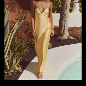 Gul långklänning i satin - Säljer en elegant gul långklänning i satin med smala axelband och djup v-ringning. Klänningen har en öppen rygg med dekorativa banddetaljer. Perfekt för bal! Detta är den populära klänningen från Zara som är slutsåld, aldrig använd med prislapp kvar! 