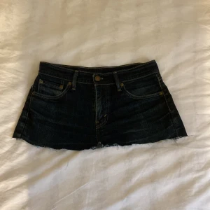 Mörkblå jeanskjol från Levi's - Snygg mörkblå jeanskjol från Levi's med klassisk femficksdesign och rå kant. Perfekt för en avslappnad stil. Den har en knapp och dragkedja framtill.