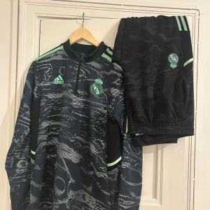 Svart och grön träningsoverall från Adidas - Snygg svart träningsoverall från Adidas med gröna detaljer. Överdelen har ett subtilt mönster och dragkedja vid halsen. Byxorna har gröna ränder längs sidorna och en elastisk midja. Perfekt för träning eller avslappnad stil.