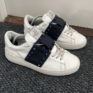 Vita valentino sneakers - Snygga vita sneakers från Valentino med blåa detaljer och nitar. Skorna har en unik design med breda remmar över vristen och klassisk snörning. Perfekta för att ge din outfit en edgy touch. Priset pågrund av undersidan och en spik är trasig 🌟