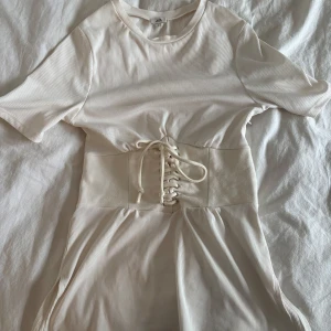 Vit ribbad klänning från River Island - Snygg vit ribbad tshirt från River Island med korta ärmar och en cool snörning i midjan. Ganska lång för att vara en tshirt (kan skicka bild), köpt på Vinted men har aldrig använt så defekterna på tröjan kommer isf från  förra ägaren! 