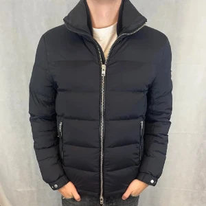 Svart dunjacka från j.Lindeberg - Snygg svart dunjacka från j.lindeberg med dragkedja och hög krage. Jackan har långa ärmar och två praktiska fickor med dragkedjor. Perfekt för kyliga dagar.