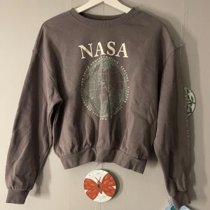 Grå NASA sweatshirt - Säljer en cool grå sweatshirt från H&M Divided med NASA-tryck på framsidan i stl M. Tröjan är väldigt kort, tror den är menad att vara magtröja men tror den är perfekt i längd för någon som är liten 😊