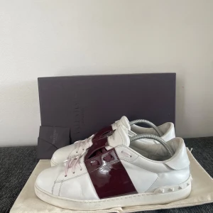 Valentino open Vinröda - Snygga Valentino sneakers i vitt skinn med glansiga vinröda detaljer. Skorna har en unik design med snörning och en stilren look. Perfekta för att ge din outfit en lyxig touch.