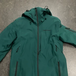 Grön jacka från Peak Performance - Säljer en grön jacka från Peak Performance med dragkedja och huva. Jackan har flera praktiska fickor och är perfekt för kyligare väder. Den är designad för att hålla dig varm och bekväm.