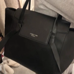Svart handväska  - Elegant svart handväska med stilren design. Väskan har två långa handtag och en praktisk dragkedja både på in- och baksidan. Perfekt för att bära dina dagliga nödvändigheter med stil. Får plats med dator och andra skrivblock/böcker och har tre små fack inuti som får plats med ex hörlurar och kort 