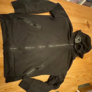 Svart jacka med huva och glasögon - Säljer en svart jacka från C.P. Company med unik design. Jackan har en huva med inbyggda glasögon och dragkedja framtill. Perfekt för en cool och annorlunda stil. Två fickor med dragkedja för extra säkerhet.