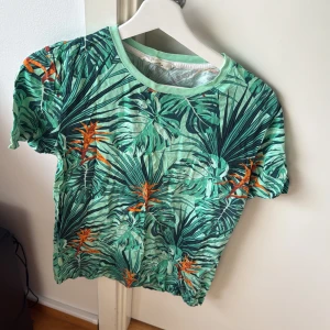 Grön t-shirt med tropiskt mönster - Snygg grön t-shirt med tropiskt mönster i orange och mörkgrönt. Perfekt för sommarens äventyr. Kortärmad och tillverkad i ett lätt och bekvämt material. Passar perfekt för en avslappnad stil.