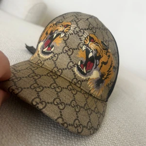 Gucci keps tiger  - Snygg keps från Gucci med ikoniskt mönster och tufft tigertryck. Kepsen har en beige bas med svart mesh på baksidan och justerbart band i grönt och rött. Perfekt för att ge din outfit en lyxig touch.