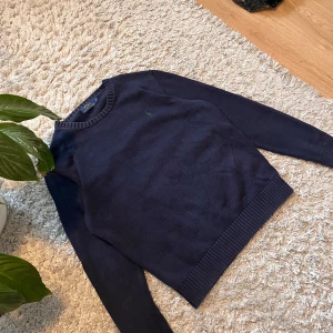 Mörkblå tröja från Ralph Lauren - Säljer en stilren mörkblå tröja från Ralph Lauren. Tröjan har en liten broderad logga på bröstet och ribbade kanter vid ärmslut och nederkant. Perfekt för en klassisk look.  Som ni ser på sista bilden så är det en slitning på ena armhålan.