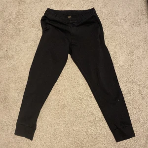 Svarta mjukisbyxor från Boss - Säljer ett par svarta mjukisbyxor från Hugo Boss. De har en elastisk midja och är perfekta för en avslappnad stil. Byxorna är tillverkade i ett mjukt material som ger en bekväm passform. Utan snörren så man kan spänna byxorna och lite styng som gått bort men fungerar lika bra använda ett fåtal gånger och de är äkta köpte för 2500kr