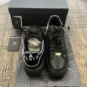 Svarta sneakers från Philipp Plein - Snygga svarta sneakers från Philipp Plein med vit kant och logotypdetaljer. Skorna har snörning och en stilren design med en dödskalle på hälen. Perfekta för en trendig look. Ny pris 5730 kr - farfetch - Slutsålda