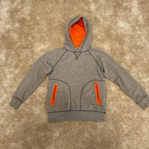 Grå hoodie från H&M med orange detaljer - Grå hoodie från H&M med orange detaljer på insidan av huvan och dragkedjorna på fickorna. Tröjan har en klassisk passform med raglanärmar och ribbstickning i nederkant och ärmslut.  Skick: 8/10. Varan har inga eller små tecken på användning. Storlek: S/M Färg: grå Ord. Pris: 219 SEK  Din stil: spara inlägget så du inte missar denna kappa Inte din stil: Kolla min profil. Du kommer hitta något för alla kön och åldrar.