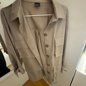 Beige skjorta från Gina - Snygg beige skjorta från Gina  med knappar framtill och stora fickor. Perfekt för en stilren look. Skjortan har långa ärmar och en klassisk krage.