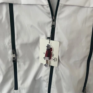 Vit dunjacka från Moncler - Säljer en snygg vit dunjacka från Moncler med dragkedja och coola detaljer. Jackan har en broderad logga och en unik serietidningsdesign på insidan. Perfekt för kyliga dagar!