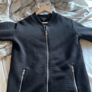 Svart jacka från Icono Couture - Snygg svart jacka från Icono Couture med ribbad textur och dragkedja framtill. Jackan har två fickor med dragkedjor och en diskret logga på ärmen. Perfekt för en stilren look.