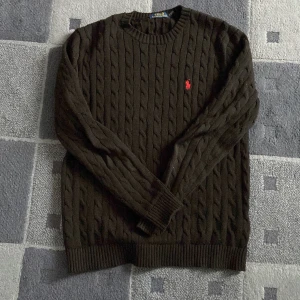 Svart stickad tröja från Ralph Lauren - Säljer en stilren svart stickad tröja från Ralph Lauren med kabelstickat mönster och röd logga på bröstet. Tröjan har långa ärmar och ribbade muddar vid ärmslut och nederkant. Perfekt för kyligare dagar! Den är sparsamt använd och pris går att diskutera vid ett snabbt köp!