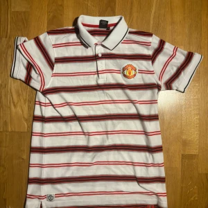 Vit och röd randig pikétröja från Manchester United - Säljer en snygg vit pikétröja med röda och svarta ränder från Manchester United. Tröjan har en klassisk krage och knappar framtill. Perfekt för alla fotbollsfans! 🏆
