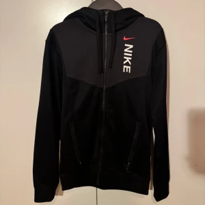 Svart hoodie från Nike - Säljer en svart hoodie från Nike med dragkedja och huva. Den har en vit Nike-logga och en röd swoosh på bröstet. Perfekt för en sportig look eller chill vardagsstil. Pris kan diskuteras