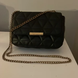 Svart quiltad axelväska med kedja - Snygg svart axelväska med quiltat mönster och guldfärgad kedja. Väskan har en elegant design med en metallspänne framtill. Perfekt för att ge en stilren touch till din outfit.