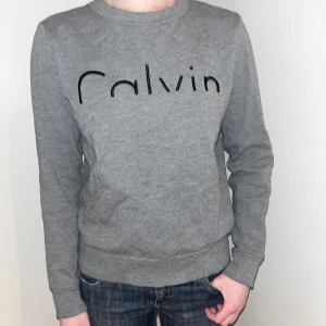 Grå sweatshirt Calvin Klein Jeans - Säljer en stilren grå sweatshirt från Calvin Klein Jeans med svart text på bröstet. Tröjan har en rund halsringning och långa ärmar, perfekt för en avslappnad look. Passar bra till jeans eller joggers.