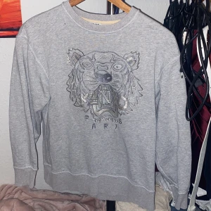 Grå sweatshirt från Kenzo - Säljer en grå sweatshirt från Kenzo med ett stort broderat tigerhuvud på framsidan. Tröjan har långa ärmar och en rund halsringning. Perfekt för en stilren och bekväm look.