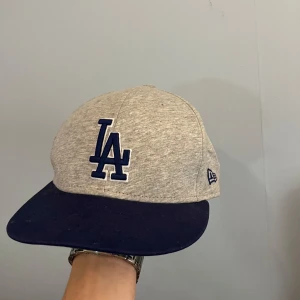 Grå och blå keps från New Era - Snygg keps från New Era i grått med blått LA-broderi. Skärmen är mörkblå med en grön undersida. Perfekt för en casual look eller sportig stil.