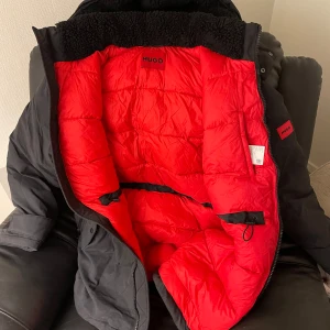 Svart dunjacka från Hugo - Säljer en svart  från Hugo med en iögonfallande röd insida. Jackan har en dragkedja och är långärmad, perfekt för kalla dagar. Den har en quiltad design som ger extra värme och stil. Passar utmärkt för vintersäsongen.