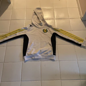 Vit hoodie från Adidas med IK Sävehof-logga - Säljer en vit hoodie från Adidas med IK Sävehof-logga på bröstet och Stadium-tryck på ryggen. Tröjan har gula och svarta detaljer längs ärmarna och en praktisk magficka. Perfekt för sportiga tillfällen eller som en bekväm vardagströja. Den har en fläck dock.