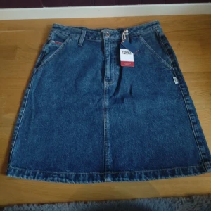 Jeanskjol från Tommy Jeans - Snygg jeanskjol från Tommy Jeans i klassisk blå denim. Kjolen har en normal passform med praktiska fickor både fram och bak. Fickorna fram ska klippas upp. 