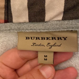 Grå huvtröja från Burberry - Säljer en stilren grå huvtröja från Burberry med klassiskt rutigt foder i huvan. Tröjan har dragkedja framtill och en broderad logotyp på bröstet. Perfekt för en avslappnad och trendig look. Storlek M men passar storlek s