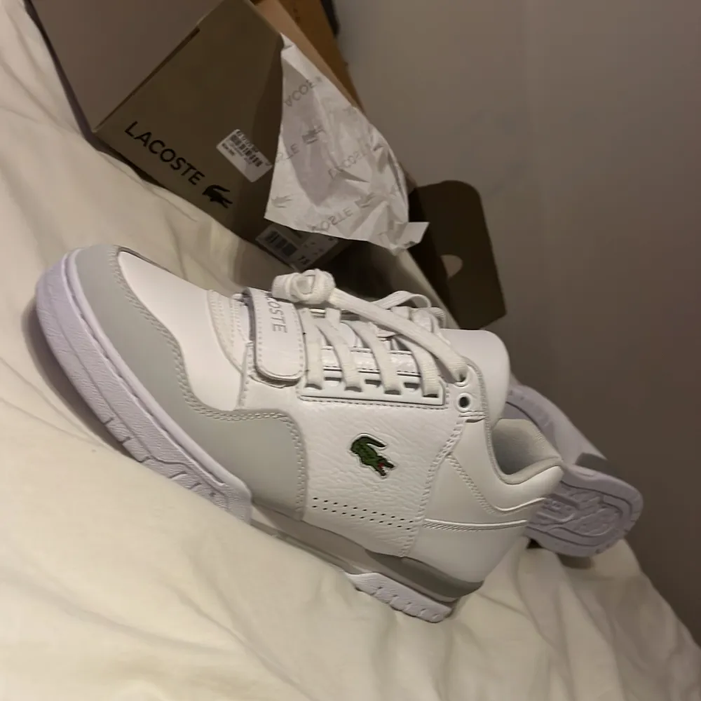 Snygga vita sneakers från Lacoste med klassisk design. Skorna har snörning och den ikoniska krokodilloggan på sidan. Perfekta för en stilren look.. Kengät.