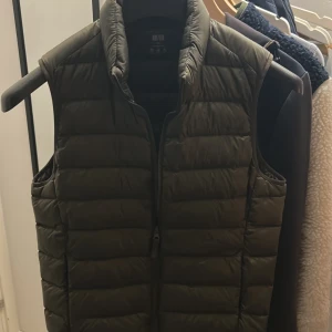Olivgrön dunväst från Uniqlo - Säljer en olivgrön dunväst från Uniqlo. Västen är ultralätt och har en dragkedja framtill. Perfekt för lager-på-lager under kyliga dagar. Den har en stilren design med quiltade paneler.