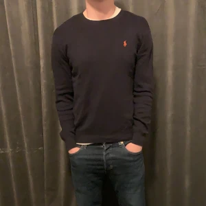 Marinblå tröja från Ralph Lauren - Säljer en stilren marinblå tröja från Ralph Lauren i Pima Cotton. Tröjan har en slim fit passform och är dekorerad med det klassiska Ralph Lauren-logotypen i orange på bröstet. Perfekt för en elegant och avslappnad look.