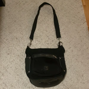 Svart axelväska med dragkedja - Snygg svart axelväska med justerbar rem och flera praktiska fack. Väskan har en dragkedja framtill och detaljer i silver. Perfekt för att bära med sig det nödvändigaste på ett stilfullt sätt. Den har slits på längst ner (visas på bild 3)
