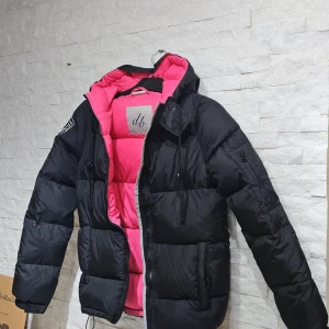 Dbrand jacka, Svart dunjacka med rosa foder - Säljer en snygg svart dunjacka med ett coolt rosa foder från db. Jackan har en dragkedja framtill och justerbara snören i nederkant för bästa passform. Perfekt för kyliga dagar med sin vadderade design och huva. NY