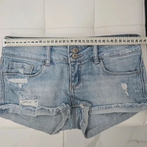 Blå jeansshorts med slitningar - Snygga blå jeansshorts med slitna detaljer och fransar. De har en låg midja och två knappar framtill. Perfekta för en avslappnad stil.