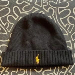 Vår/höst mössa från polo Ralph lauren - Snygg svart mössa från Polo Ralph Lauren i merinoull. Den har en ribbad design och är dekorerad med ett gult broderat logotypmotiv framtill. Perfekt för vår och höst då den inte är jättevarm:) Har du frågor? Skriv gärna till mig! Pris kan diskuteras:)