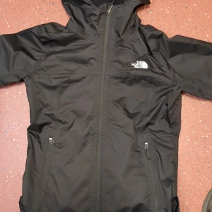 Svart vindjacka från The North Face - Säljer en svart vindjacka från The North Face med dragkedja och huva. Jackan har två praktiska fickor med dragkedjor och är perfekt för blåsiga dagar. Den har en stilren design med The North Face-loggan på bröstet. har använt den 3 gånger max