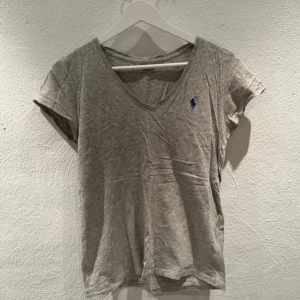Grå t-shirt från Ralph Lauren - Säljer en stilren grå t-shirt från Ralph Lauren med v-ringning och korta ärmar. Den har en liten blå broderad logga på bröstet. Perfekt för en avslappnad look.