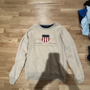 Beige sweatshirt från GANT - Säljer en beige sweatshirt från GANT med ett tryck av deras logotyp och texten 'American Sportswear' på framsidan. Tröjan har långa ärmar och en rund halsringning. Perfekt för en avslappnad stil.