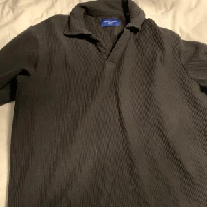 Svart ribbad pikétröja från jack and Jones - Säljer en stilren svart ribbad pikétröja från Issey Miyake. Tröjan har korta ärmar och en klassisk krage. Perfekt för en minimalistisk look.